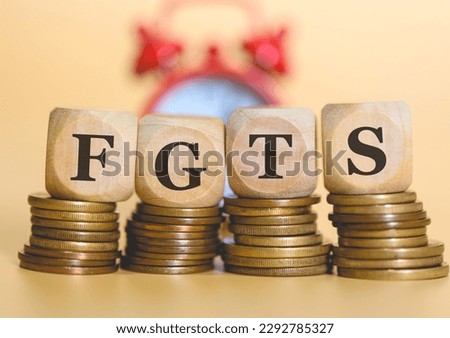The acronym FGTS written on wooden dice lying on piles of coins in a studio photo. An alarm clock in the background in the composition. Brazilian economy.
