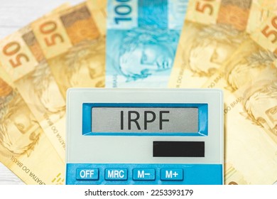 The acronym IRPF referring to the Personal Income Tax in Brazil in Brazilian Portuguese written on the display of a calculator with banknotes of reais in the composition. Brazilian economy and investm - Shutterstock ID 2253393179