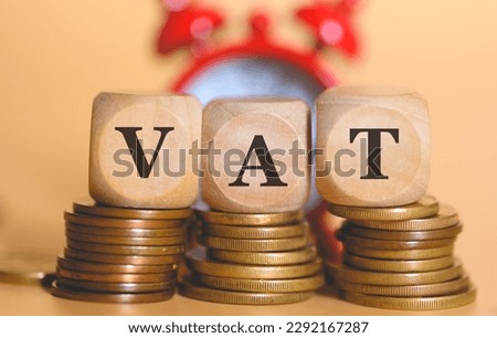 The acronym VAT for Value-Added Tax written on wooden dice lying on top of piles of coins. Studio photo. An alarm clock in the composition.
