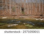 Across the flowing whitewater creek with boulders and fallen trees on the shoreline looking at the water trickling down the stacked rock bluff surrounded by the bare tree forest in wintertime