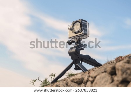 An action camera in a protective case mounted on a mini tripod stands on a rocky surface with a clear blue sky. Ideal for shooting outdoor adventures and nature photos.