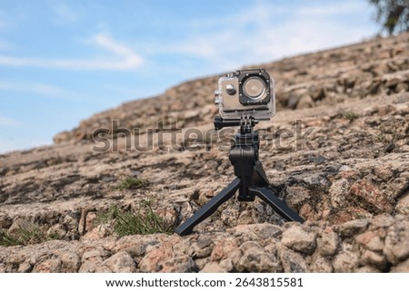 An action camera in a protective case mounted on a mini tripod stands on a rocky surface with a clear blue sky. Ideal for shooting outdoor adventures and nature photos.