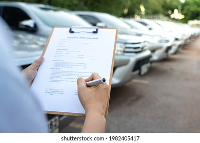 Action of a customer is signing on the agreement term of car rental service. Close-up and selective focus a human's hand with blurred background of cars in row. Business and transportation concept.