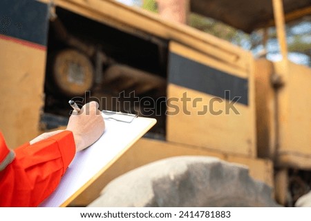 Action of an engineer hand is writing to perform inspection of the road roller or road compactor machinery engine part (as blurred background). Industrial working action scene, selective focus. 