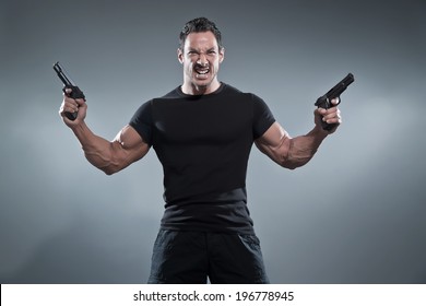 Action Hero Muscled Man Holding Two Guns. Wearing Black T-shirt And Pants. Studio Shot Against Grey.