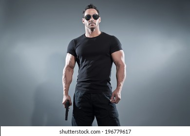 Action hero muscled man holding a gun. Wearing black t-shirt with pants and sunglasses. Studio shot against grey.