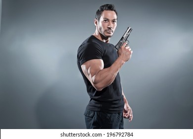 Action hero muscled man holding a gun. Wearing black t-shirt and pants. Studio shot against grey.