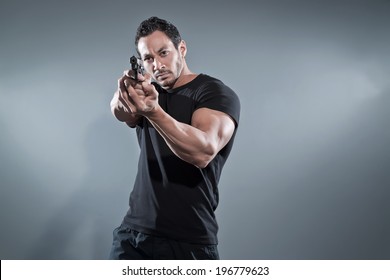 Action Hero Muscled Man Shooting With Gun. Wearing Black T-shirt And Pants. Studio Shot Against Grey.