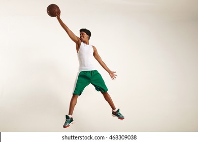 Action photo of a happy young black athlete wearing white shirt and green shorts jumping high to grab a vintage basketball against white background