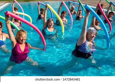 Action portrait of senior aqua gym class. Elderly ladies exercising together with foam noodles in outdoor swimming pool.
