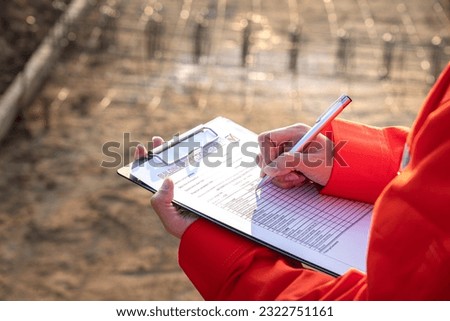 Action of a safety engineer is checking the building quality report form with construction work site as background. Industrial working action scene with people, close-up and selective focus.