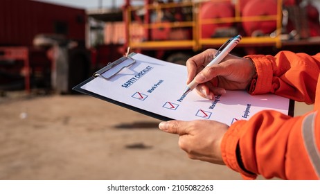 Action Of Safety Office Is Writing On Checklist Paper During Safety Audit And Risk Verification At Drilling Site Operation With Blurred Background Of Mount Truck Rig. Selective Focus At Hand.