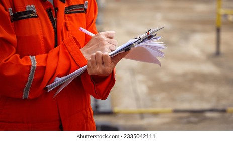 Action of a safety officer in full PPE coverall is writing note on paper document during perform safety audit at construction worksite. Industrial expert working scene. Selective focus.