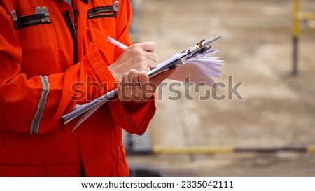 Action of a safety officer in full PPE coverall is writing note on paper document during perform safety audit at construction worksite. Industrial expert working scene. Selective focus.
