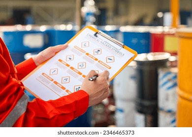 Action of safety officer is using a pen to checking on the hazadous material symbol label form with the chemical barrel as blurred background. Safety industrial working scene concept. Selective focus.