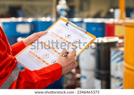Action of safety officer is using a pen to checking on the hazadous material symbol label form with the chemical barrel as blurred background. Safety industrial working scene concept. Selective focus.