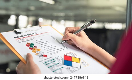 Action of a safety supervisor hand is checking on checklist of chemical and hazardous material form to verify the risk with blurred background of factory storage workplace. Industrial working scene.