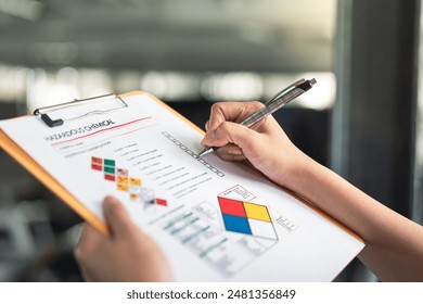 Action of a safety supervisor hand is checking on checklist of chemical and hazardous material form to verify the risk with blurred background of factory storage workplace. Industrial working scene.