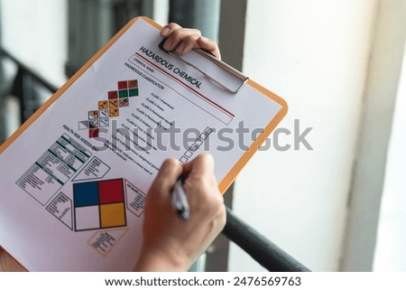 Action of a safety supervisor hand is checking on checklist of chemical and hazardous material form to verify the risk with blurred background of factory storage workplace. Industrial working scene.