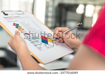 Action of a safety supervisor hand is checking on checklist of chemical and hazardous material form to verify the risk with blurred background of factory storage workplace. Industrial working scene.