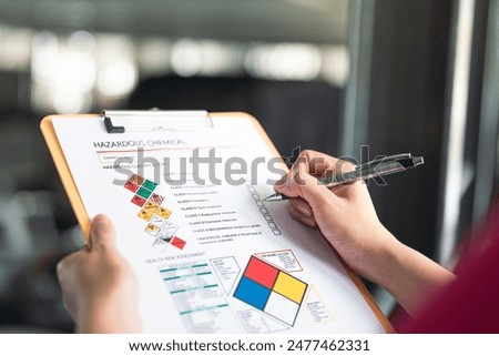 Action of a safety supervisor hand is checking on checklist of chemical and hazardous material form to verify the risk with blurred background of factory storage workplace. Industrial working scene.