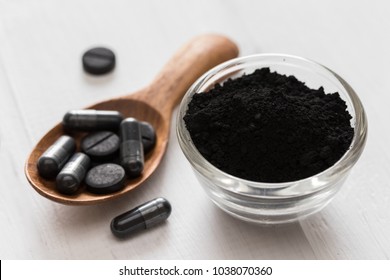Activated charcoal powder in a glass bowl and pills on a spoon on white wooden background