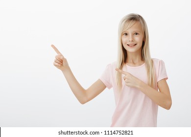 Active Adorable Female Child With Blond Hair In Trendy Pink T-shirt, Pointing At Upper Left Corner And Smiling Broadly, Asking Parents Permission To Ride On Ferris Wheel, Standing Over Gray Background