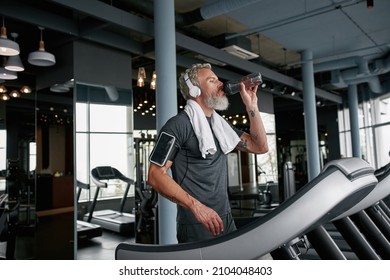 Active aged bearded man with towel around neck drinking water from bottle standing on treadmill in gym. Mature man in headphones after running in gym. Healthy lifestyle. - Powered by Shutterstock