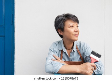 Active Asian Mature Woman Sitting Outdoor With A Red Brush On Her Hand For Painting. Senior Woman Relaxing On A Chair In Front Of White Wall And Blue Door. Portrait Of Middle Aged Woman Smiling.