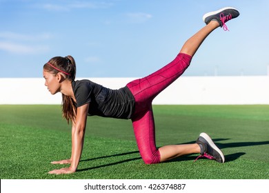 Active Asian young adult doing bodyweight glute and leg exercises on outdoor grass. Fitness woman doing donkey kick exercise for glutes strength training, butt toning and body core health.