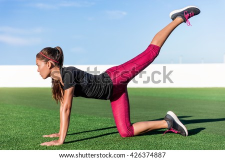 Active Asian young adult doing bodyweight glute and leg exercises on outdoor grass. Fitness woman doing donkey kick exercise for glutes strength training, butt toning and body core health.
