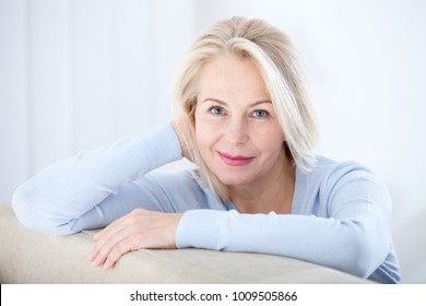 Active beautiful middle-aged woman smiling friendly and looking in camera in living room. Woman's face closeup. Realistic images without retouching with their own imperfections. Selective focus.