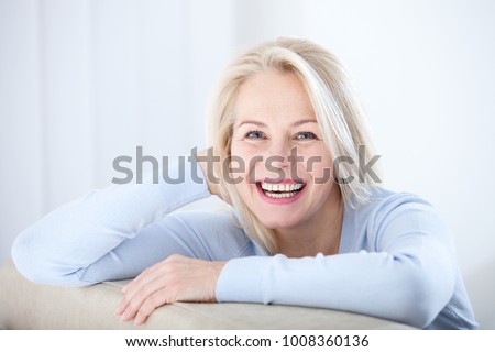 Active beautiful middle-aged woman smiling friendly and looking in camera in living room. Woman's face closeup. Realistic images without retouching with their own imperfections. Selective focus.