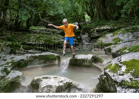 Active boy jumps from rock to rock on a calm mountain river hidden in a dense forest
