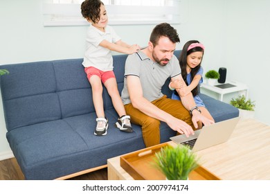 Active children with a lot of energy bothering their working dad. Hispanic man trying to work and type on the laptop while his kids are trying to play with him