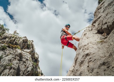 Active climber middle age man in protective helmet looking at camera while abseiling from cliff rock wall using rope with belay device and climbing harness. Active extreme sports time spending concept