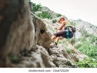 Active climber woman in protective helmet abseiling from cliff rock wall using a rope with a belay device and climbing harness. Active extreme sports time spending concept.