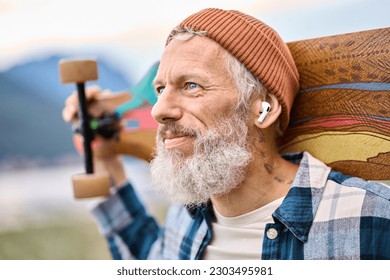 Active cool bearded old hipster man standing in nature park holding skateboard wearing earbud. Mature traveler skater enjoying freedom spirit and extreme sports hobby listening music in earphones.
