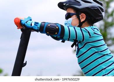 Active Cute Boy Wearing Protective Mask And Gear Playing And Riding Sport Scooter At Park. Kids Summer Activities Background Under Covid-19 Pandemic New Normal.