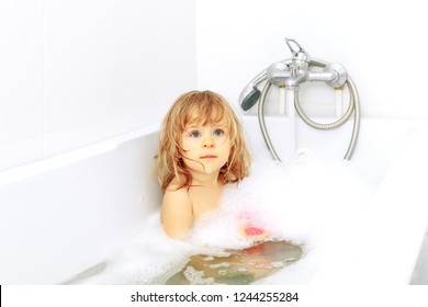 Active cute little blond toddler girl of 2 years having fun with water by taking bath in bathtub at home. Funny kid splashing and playing with toys. Sad baby in the shower.
