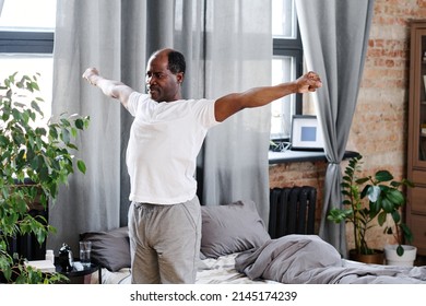 Active Elderly Black Man With Outstretched Arms Exercising In The Morning While Standing By His Bed With Grey Pillows And Blanket