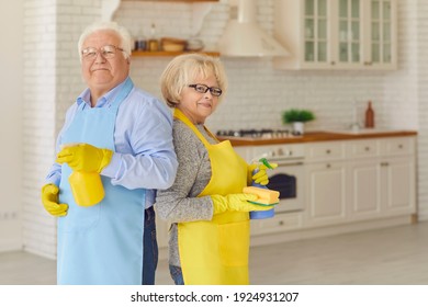 Active Elderly Couple Holding Cleaning Detergents And Standing With Their Backs To Each Other In The Kitchen In Aprons And Gloves. Concept Of Housework, Cleanliness, Tidiness In Older People.