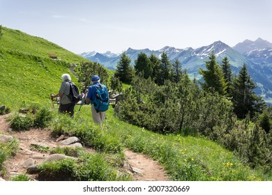 An Active Elderly Couple (no Faces Visible) With Backpacks And Special Sticks Are Hiking In The Mountains And Enjoying Beautiful Views Of The Alps. Retirement Healthy And Active Lifestyle Concept.