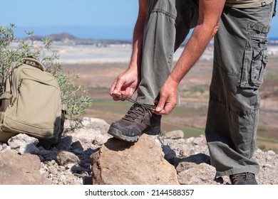 Active Elderly Man Hiking Outdoors In The Mountains Tying His Shoes Enjoying Healthy Lifestyle And Sunny Day. Panoramic View Of The Sea And The Mountain In Background