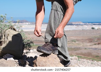 Active Elderly Man Hiking Outdoors In The Mountains Tying His Shoes Enjoying Healthy Lifestyle And Sunny Day. Panoramic View Of The Sea And The Mountain In Background