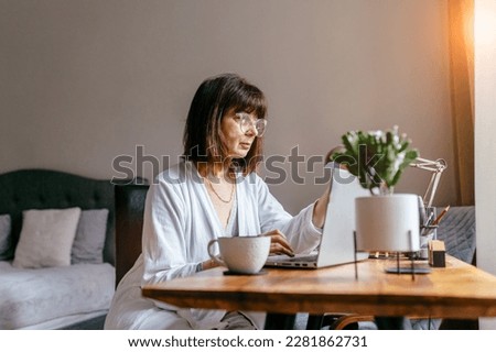 Active female senior in a bathrobe working with laptop in bedroom enjoying retirement with leisurely daily routine. Make research at home. Jotting information, create to-do list, planning concept