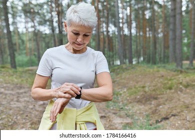 Active Fit Retired Female Runner Taking Break During Morning Run Looking At Her Wrist Watch. Serious Mature Woman Athlete Setting Up Fitness Tracker To Monitor Heart Rate While Running In Park