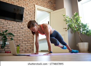 Active fit sporty young woman wearing sportswear doing fitness plank exercise in living room at home on mat watching online video training tutorial sport live stream workout class on laptop computer.
