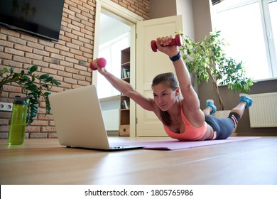 Active fit sporty young woman holding dumbbells doing fitness exercise training abs and back at home on mat watching online video training tv tutorial sport live stream workout class on laptop.