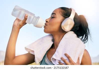 An Active Fit Woman Wearing Wireless Headphones Drinking Water From A Bottle After Exercising Outdoors. Female Athlete Quenching Thirst And Cooling Down With A Towel After Training Workout Outside